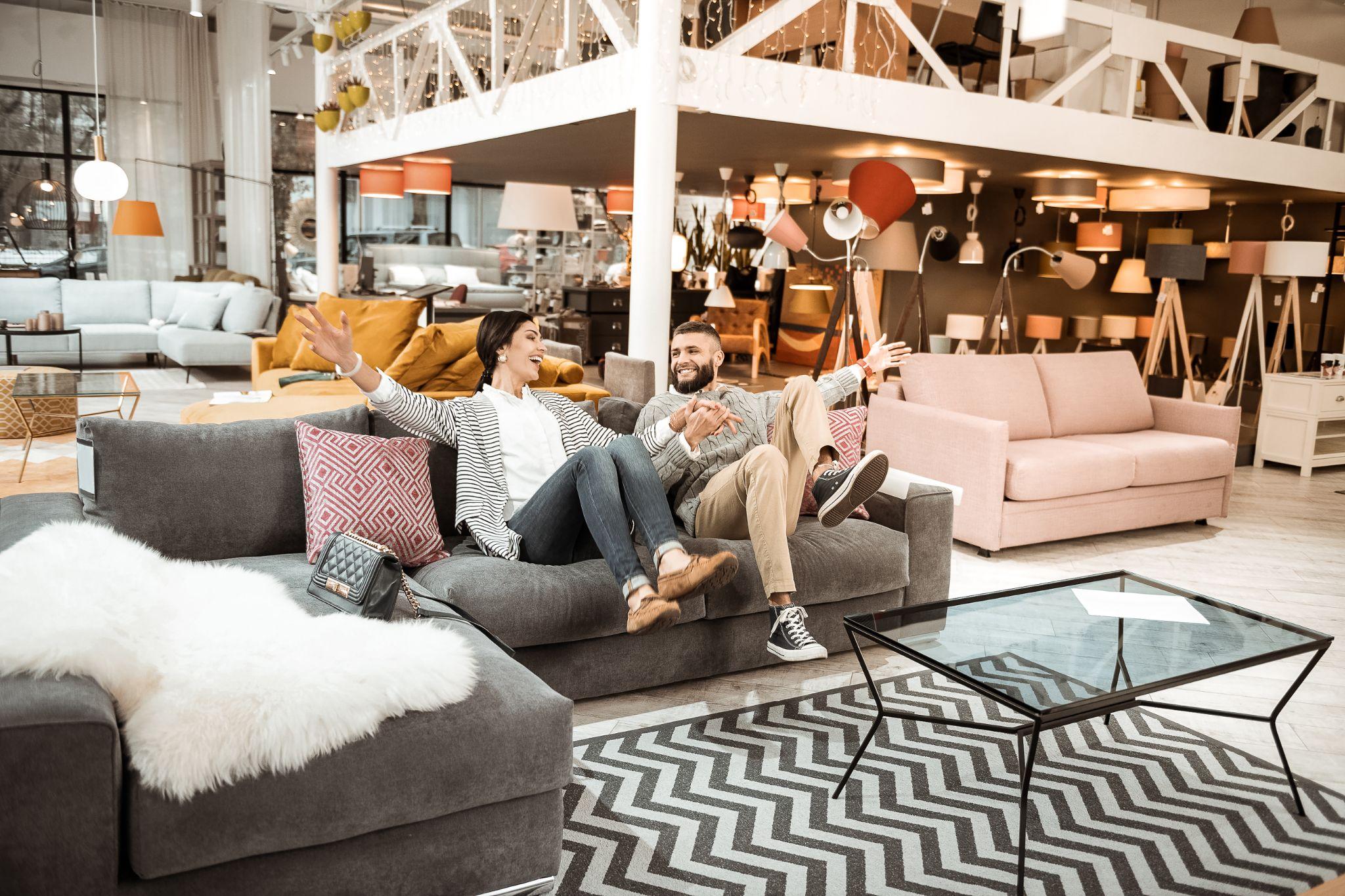 Cheerful laughing couple behaving childishly in furniture showroom while fooling around on grey couch